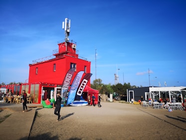A vibrant red building with a rooftop antenna in a sandy area is surrounded by several people dressed in casual or sporty attire. There are large banners in front of the building, and a dining area with outdoor seating on the right. The sky is clear and blue, indicating a sunny day.