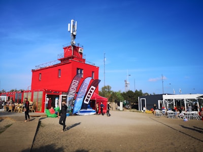 A vibrant red building with a rooftop antenna in a sandy area is surrounded by several people dressed in casual or sporty attire. There are large banners in front of the building, and a dining area with outdoor seating on the right. The sky is clear and blue, indicating a sunny day.