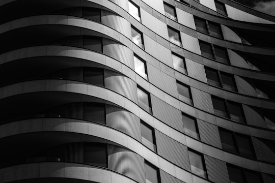 A black and white architectural photograph featuring a modern building with a curved facade. The facade has a repetitive pattern of alternating balconies and rectangular windows, creating a sense of rhythm and movement. The shadows and light create a dramatic contrast, highlighting the texture and design of the building.