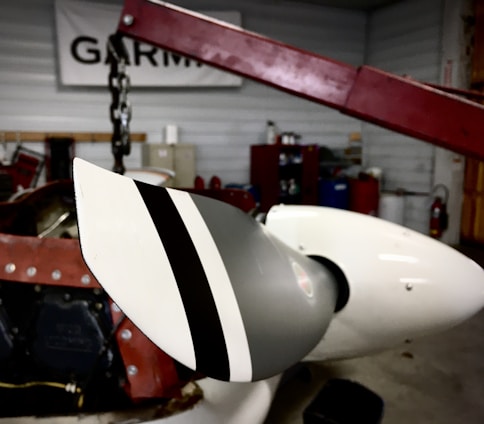 A close-up of a propeller blade attached to an aircraft engine. The blade has a distinctive black and white striped pattern, and the background features an indoor workshop setting with tools, equipment, and a sign displaying the word 'GARMIN'.