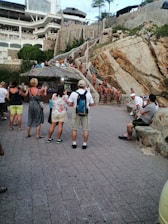 A gathering of people near a rugged cliffside, with multiple individuals in swimsuits standing on stairs and natural rock formations. Tourists observe and take pictures, some are under a thatched roof structure. The scene includes a large building in the background with balconies, surrounded by lush greenery.
