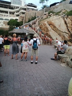 A gathering of people near a rugged cliffside, with multiple individuals in swimsuits standing on stairs and natural rock formations. Tourists observe and take pictures, some are under a thatched roof structure. The scene includes a large building in the background with balconies, surrounded by lush greenery.