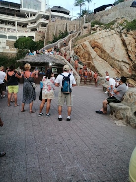 A gathering of people near a rugged cliffside, with multiple individuals in swimsuits standing on stairs and natural rock formations. Tourists observe and take pictures, some are under a thatched roof structure. The scene includes a large building in the background with balconies, surrounded by lush greenery.