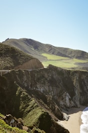 A coastal landscape featuring rugged cliffs and green hills under a clear blue sky. The shoreline comprises rocky cliffs that descend sharply into a sandy beach below. The vegetation on the hills appears lush and dense, indicating a vibrant natural setting.