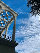 A geometric roof structure is supported by tall columns against a clear blue sky, partially filled with light, fluffy clouds. A tree with dark green needles is visible in the top right corner, adding a natural element to the architectural scene.
