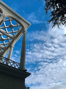 A geometric roof structure is supported by tall columns against a clear blue sky, partially filled with light, fluffy clouds. A tree with dark green needles is visible in the top right corner, adding a natural element to the architectural scene.