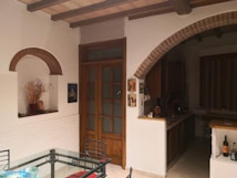 A cozy indoor space featuring a dining area with a glass-top table and metal chairs. The room has a rustic aesthetic with wooden beams and arched brick details. A wooden door with frosted glass panels is situated centrally. The kitchen area is visible with countertops and cupboards, and various decorative items are placed throughout, including a vase with dried plants and wall art.