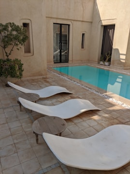A serene courtyard featuring a small swimming pool surrounded by a tiled patio. Modern, curved lounge chairs are arranged next to the pool with a small round wooden table beside them. The walls are made of beige stucco, and large glass doors lead to an interior space. A small tree and potted plants add a touch of greenery to the minimalist design.