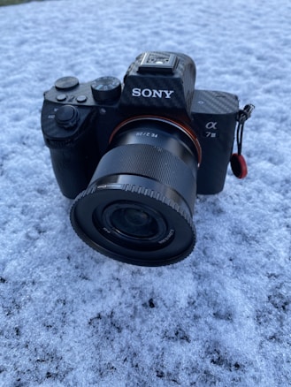 A black Sony camera with a lens is placed on a snowy surface. The camera appears to be a mirrorless model from the Alpha series, featuring a textured grip and various control dials. The lens is mounted securely, and some of the camera's branding details are visible.
