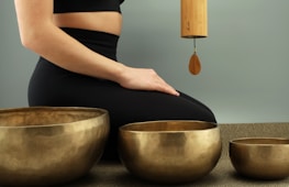 A person is seated in a calm, meditative posture on a mat, with three golden singing bowls placed in front of them. A wind chime made of bamboo is suspended, adding a serene ambiance to the scene. The individual is dressed in black athletic wear.