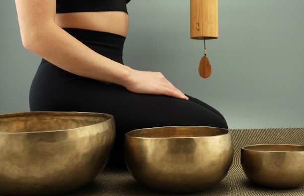 A person is seated in a calm, meditative posture on a mat, with three golden singing bowls placed in front of them. A wind chime made of bamboo is suspended, adding a serene ambiance to the scene. The individual is dressed in black athletic wear.
