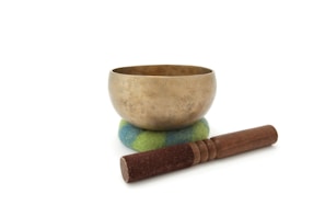 A brass singing bowl sits on a colorful cushion with blue and green hues. In front of the bowl, there is a wooden mallet with a suede-wrapped end.