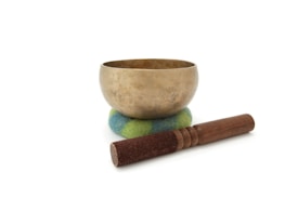 A brass singing bowl sits on a colorful cushion with blue and green hues. In front of the bowl, there is a wooden mallet with a suede-wrapped end.