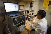A person wearing a lab coat and safety goggles is using a microscope at a laboratory workstation. A laptop and various scientific instruments are visible on the table, displaying data and metrics.