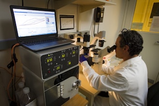 A person wearing a lab coat and safety goggles is using a microscope at a laboratory workstation. A laptop and various scientific instruments are visible on the table, displaying data and metrics.