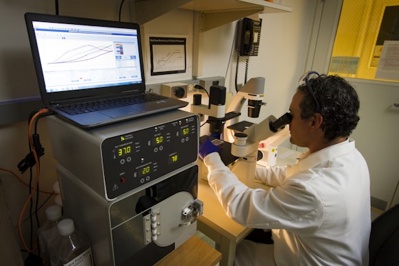 A person wearing a lab coat and safety goggles is using a microscope at a laboratory workstation. A laptop and various scientific instruments are visible on the table, displaying data and metrics.