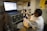 A person wearing a lab coat and safety goggles is using a microscope at a laboratory workstation. A laptop and various scientific instruments are visible on the table, displaying data and metrics.