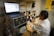 A person wearing a lab coat and safety goggles is using a microscope at a laboratory workstation. A laptop and various scientific instruments are visible on the table, displaying data and metrics.