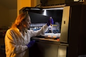 A person wearing protective gloves and a lab coat is working with a pipette inside a laboratory. They are transferring liquid into a container and focused on their task. The environment has scientific equipment and containers labeled with various substances.