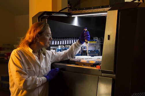 A person wearing protective gloves and a lab coat is working with a pipette inside a laboratory. They are transferring liquid into a container and focused on their task. The environment has scientific equipment and containers labeled with various substances.