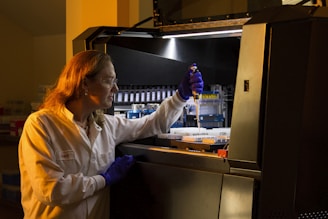 A person wearing protective gloves and a lab coat is working with a pipette inside a laboratory. They are transferring liquid into a container and focused on their task. The environment has scientific equipment and containers labeled with various substances.