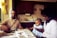 A family scene in a kitchen with a man, woman, and a small child engaging with a toy car. The man is tinkering with parts laid out on the table, which is covered with newspapers or a magazine. The woman and child, who is sitting on her lap, are observing. The kitchen features dark wooden cabinets and is softly lit by natural light coming through a window with yellow curtains.