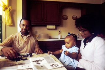 A family scene in a kitchen with a man, woman, and a small child engaging with a toy car. The man is tinkering with parts laid out on the table, which is covered with newspapers or a magazine. The woman and child, who is sitting on her lap, are observing. The kitchen features dark wooden cabinets and is softly lit by natural light coming through a window with yellow curtains.