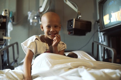 A child with a bald head is smiling while sitting on a hospital bed. The child is wearing a hospital gown and is surrounded by medical equipment, including a television and overhead monitors.