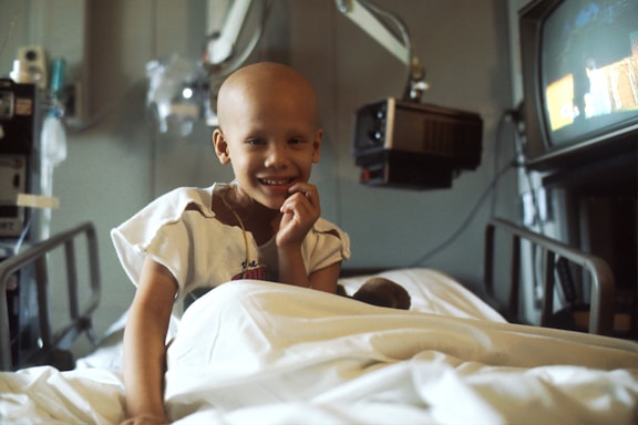 A child with a bald head is smiling while sitting on a hospital bed. The child is wearing a hospital gown and is surrounded by medical equipment, including a television and overhead monitors.