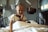 A child with a bald head is smiling while sitting on a hospital bed. The child is wearing a hospital gown and is surrounded by medical equipment, including a television and overhead monitors.