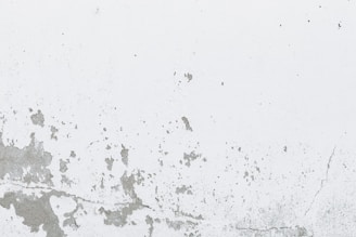 A white wall with peeling and cracking paint, showing signs of aging and wear. The texture of the surface is rough, with various patches and cracks forming irregular patterns.