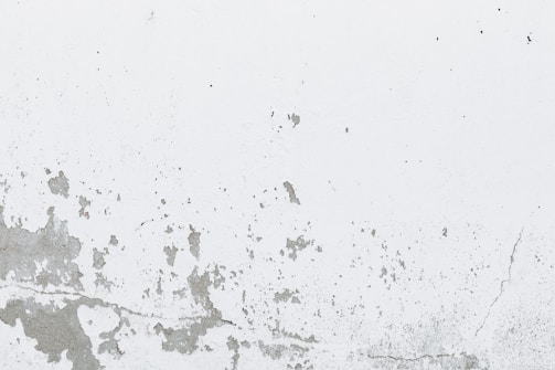 A white wall with peeling and cracking paint, showing signs of aging and wear. The texture of the surface is rough, with various patches and cracks forming irregular patterns.