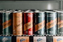 A row of assorted Schweppes canned beverages is arranged on a shelf. The cans feature various flavors including Dry Lemon, Tonic Water, Ginger Ale, and Lemonade. They display vibrant labels with prominent branding in a vertical refrigerator.