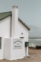 A small, white building with a steep green roof and a chimney sits near the coast. The building is marked with a sign that reads 'Strandveld Museum.' The background features a cloudy sky and a view of the ocean, with waves gently breaking near the shoreline.