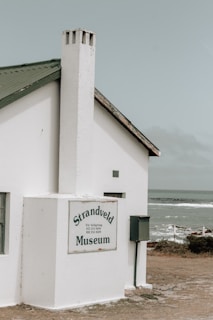 A small, white building with a steep green roof and a chimney sits near the coast. The building is marked with a sign that reads 'Strandveld Museum.' The background features a cloudy sky and a view of the ocean, with waves gently breaking near the shoreline.