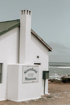 A small, white building with a steep green roof and a chimney sits near the coast. The building is marked with a sign that reads 'Strandveld Museum.' The background features a cloudy sky and a view of the ocean, with waves gently breaking near the shoreline.