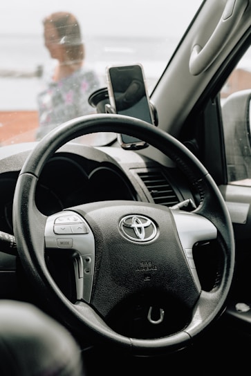 A steering wheel with the Toyota logo is prominently featured inside a car. A smartphone is mounted on the dashboard in a holder. Outside the car window, a blurry human figure is visible in the background.