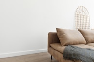A beige sofa with plush cushions is positioned against a white wall. A decorative wooden lattice is leaning against the wall, and a gray textured blanket is draped over the arm of the sofa. The room features a wooden floor, adding a warm and minimalist aesthetic.