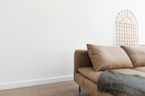 A beige sofa with plush cushions is positioned against a white wall. A decorative wooden lattice is leaning against the wall, and a gray textured blanket is draped over the arm of the sofa. The room features a wooden floor, adding a warm and minimalist aesthetic.