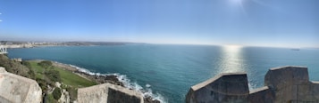 A panoramic view of a vast, calm ocean meeting the sky at the horizon, with sunlight reflecting on the water. Foreground features a historic stone parapet overlooking coastal greenery and rocky shores. In the distance, a ship is visible on the water and a coastline stretches into the horizon.