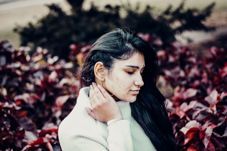 A woman with long dark hair stands among lush, maroon-colored foliage, wearing a light-colored knit sweater. She is holding her neck gently with one hand, and her eyes are closed, conveying a sense of calmness or introspection.