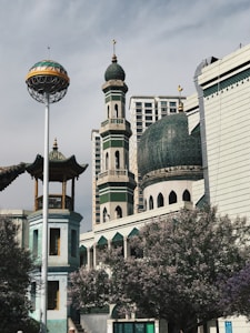An architectural scene featuring a mosque with a large green dome and a tall minaret. The mosque is adorned with detailed patterns and is situated in an urban setting with modern buildings in the background. There is a flowering tree in the foreground with pink blossoms and a metal globe structure on a tall pole nearby.
