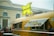 A vintage-style food truck painted yellow with colorful illustrations is parked near a building with arched windows. A large retro-style sign with a yellow 'X' and the word 'BURGERS' is displayed on top of the vehicle. People are lined up in warm clothing, suggesting cold weather. The truck's side window is open, indicating it is serving customers.