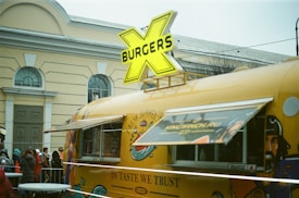 A vintage-style food truck painted yellow with colorful illustrations is parked near a building with arched windows. A large retro-style sign with a yellow 'X' and the word 'BURGERS' is displayed on top of the vehicle. People are lined up in warm clothing, suggesting cold weather. The truck's side window is open, indicating it is serving customers.