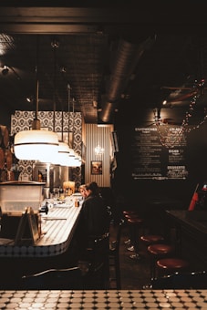 A dimly lit bar with a long counter featuring a retro design, complete with patterned tiles and hanging pendant lights. Two people are sitting at the bar, engaging in conversation. On the wall behind the bar, there are various black-and-white patterns and a menu written in chalk. The atmosphere is cozy, with warm lighting enhancing the intimate setting.