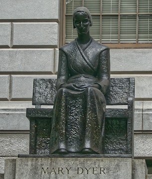 A dark metal statue of a seated woman wearing a long dress and bonnet is positioned against a textured stone wall. Her hands are folded in her lap and she has an expression of calm seriousness. The pedestal below includes an inscription of 'Mary Dyer.'