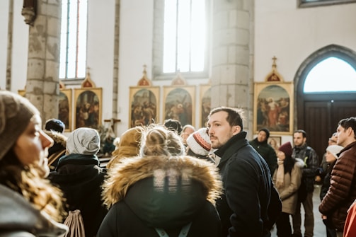 A group of people stand inside a church, surrounded by religious artwork and decorative columns. They appear to be engaged in a group tour or gathering. The light streaming through the windows casts a warm glow on the scene.