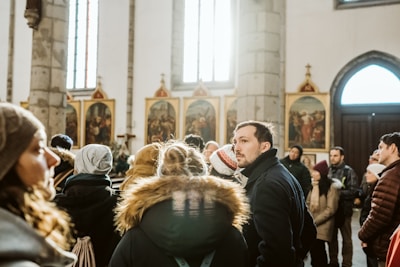 A group of people stand inside a church, surrounded by religious artwork and decorative columns. They appear to be engaged in a group tour or gathering. The light streaming through the windows casts a warm glow on the scene.