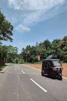 A rural road curves gently, bordered by lush green trees and a bright blue sky. A black tuk-tuk is traveling along the road. A red road sign is visible on the right, and palm trees are scattered in the background.