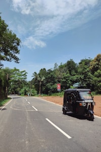 A rural road curves gently, bordered by lush green trees and a bright blue sky. A black tuk-tuk is traveling along the road. A red road sign is visible on the right, and palm trees are scattered in the background.